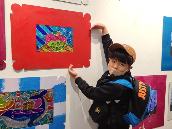 미술전시회에 참가한 온누리M센터 지역아동센터 학생이 자신이 그린 하트 그림에 하트 모양을 하며 사진 촬영을 진행하고 있다(사진 = 김종현 기자)